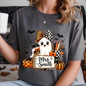 Może przedstawiać: Ciemnoszary t-shirt z grafiką przedstawiającą białego ducha w kokardzie w panterkę i kratkę, trzymającego kubek kawy. Wz&oacute;r zawiera dynie, oł&oacute;wki w kubku w kratkę, pajęczynę, nietoperze oraz notatnik z napisem Mrs. Smith.