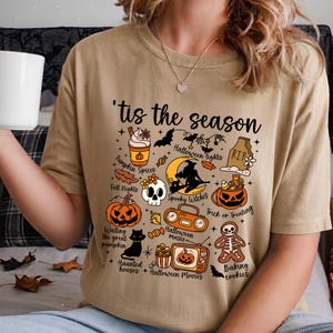 May include: A beige t-shirt featuring the text 'tis the season and various Halloween-themed illustrations including a pumpkin spice drink, a skull, a black cat, a jack-o'-lantern, a witch on a moon, a boombox, a television, and a skeleton cookie.