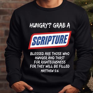 May include: A black long-sleeve sweatshirt featuring white text that reads HUNGRY? GRAB A, followed by a red, white, and blue logo graphic with the word SCRIPTURE, and further text reading BLESSED ARE THOSE WHO HUNGER AND THIRST FOR RIGHTEOUSNESS FOR THEY WILL BE FILLED MATTHEW 5:6.