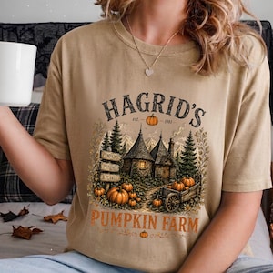 May include: A tan t-shirt featuring a graphic print of a stone cottage surrounded by pine trees and pumpkins. The text reads HAGRID'S est. 1981, PUMPKINS, POTIONS & PLANTS, and PUMPKIN FARM. A wooden signpost and a cart filled with pumpkins are visible.