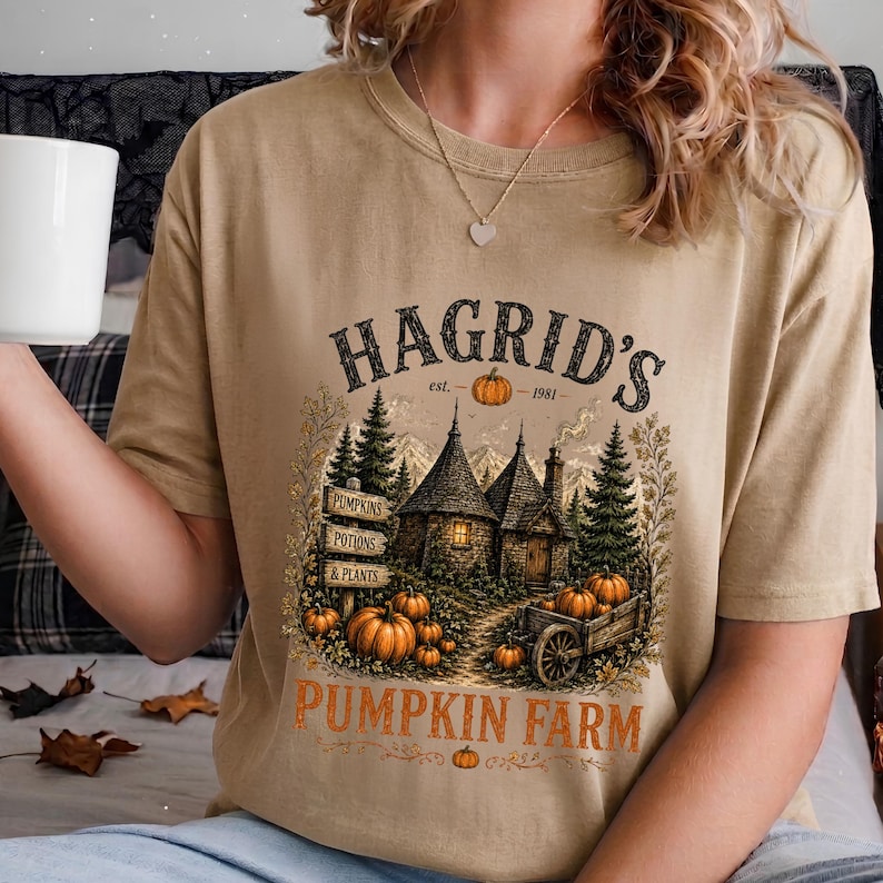 May include: A tan t-shirt featuring a graphic print of a stone cottage surrounded by pine trees and pumpkins. The text reads HAGRID'S est. 1981, PUMPKINS, POTIONS & PLANTS, and PUMPKIN FARM. A wooden signpost and a cart filled with pumpkins are visible.