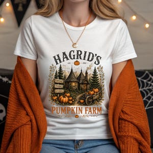 May include: A white t-shirt featuring a graphic print of a rustic cottage, pine trees, and pumpkins. The text reads HAGRID'S est. 1981 PUMPKIN FARM, with a small sign labeled PUMPKINS, POTIONS & PLANTS. The design includes a wooden cart and autumn foliage.