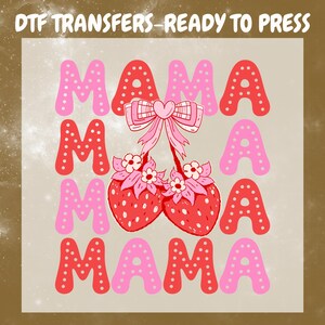 May include: A DTF transfer design featuring the word "MAMA" in pink and red bubble letters. Two red strawberries with pink flowers and a pink bow are in the center. The text "DTF TRANSFERS - READY TO PRESS" is at the top.