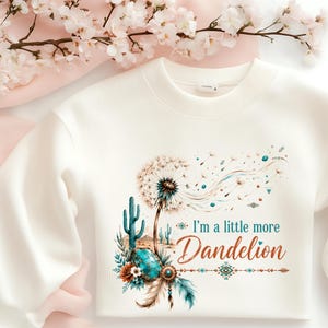 May include: Cream-colored sweatshirt featuring a dandelion design with the text "I'm a little more Dandelion." The graphic includes a cactus, turquoise accents, and feathers. The sweatshirt is a crew neck style.