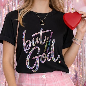 May include: A black short-sleeved t-shirt featuring the text "but God" in a multi-colored rhinestone design. Smaller text reads "He makes a way where there is no way" in pink and blue. The shirt is paired with a red heart-shaped accessory.