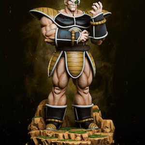 May include: A detailed figurine of a bald, muscular character in a black and gold outfit, standing on a rocky base. The figure has a green eyepiece and is posed with clenched fists. The overall design suggests a fantasy or anime theme.