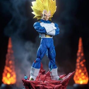 Puede incluir: Figura detallada de un personaje con pelo amarillo puntiagudo, armadura azul y blanca y brazos cruzados. La figura se alza sobre una base roja y rocosa con un soporte circular negro. El fondo es oscuro con efectos de humo y dos luces naranjas.