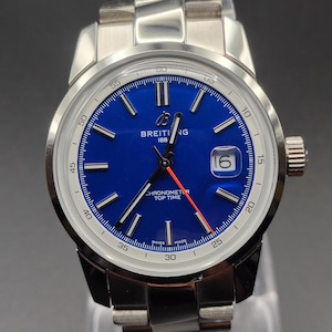 Könnte beinhalten: Eine Breitling-Armbanduhr aus Edelstahl mit einem leuchtend blauen Zifferblatt, silberfarbenen Stundenmarkierungen und einem Datumsfenster auf der Drei-Uhr-Position. Die Uhr hat einen leuchtend roten Sekundenzeiger, ein Metallgliederarmband und den Schriftzug Breitling 1884.