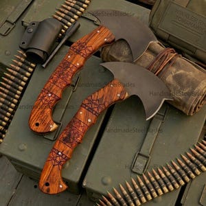 May include: Two hand axes with dark matte metal heads and curved wooden handles featuring intricate geometric laser-engraved patterns. The axes are secured with silver rivets and positioned alongside ammunition belts and a tactical pouch.