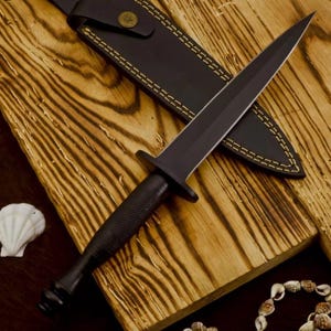 May include: A double-edged dagger with a matte black finish, featuring a textured grip and a crossguard. The dagger is accompanied by a black leather sheath with visible yellow stitching and a metallic snap button closure on the strap.
