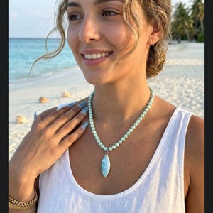 Könnte beinhalten: Eine hellgrüne Perlenkette mit Anhänger. Der Anhänger hat eine hellblaue ovale Form. Die Kette wird über einem weißen Tanktop getragen. Ein goldenes Armband befindet sich am Handgelenk.