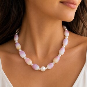 Puede incluir: Un collar con cuentas de forma ovalada en un degradado de colores rosa suave y crema, intercaladas con pequeñas cuentas esféricas blancas. El collar está centrado por una cuenta esférica blanca única y más grande en la parte inferior.