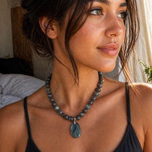 Könnte beinhalten: Eine Perlenkette mit runden, dunkelgrauen Labradoritperlen mit irisierenden blauen Reflexen. Die Kette verfügt über einen zentralen, ovalen Labradoritanhänger, der an einer silberfarbenen Metallöse befestigt ist und am Hals aufliegt.