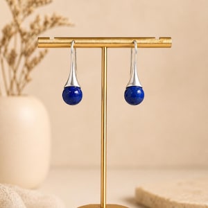 Könnte beinhalten: Ein Paar silberfarbene Ohrhänger mit Tropfendesign. Jeder Ohrring hat eine polierte silberne Kappe und einen tiefblauen, kugelförmigen Edelstein. Die Ohrringe werden auf einem goldfarbenen Schmuckständer präsentiert.