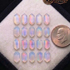 Peut inclure: Une collection de vingt pierres précieuses hexagonales iridescentes disposées en rangées dans une boîte noire. Une pièce de monnaie en argent est placée à droite pour la comparaison des tailles. Les pierres précieuses affichent une gamme de couleurs, dont le rose, le bleu et le jaune. Le logo Shifa Gems & Jewels est en haut à droite.