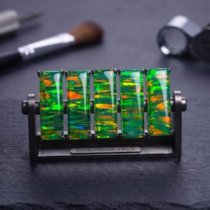 May include: Five rectangular, iridescent green and orange opal gemstones are set vertically in a dark metal frame. The base of the frame features the engraved text SHIFAGEMSANDJEWELS. The stones display a vibrant play of colour with flashes of neon green and bright orange throughout their polished surfaces.