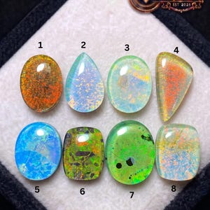 May include: Eight opals of different shapes and colours are displayed, including oval, teardrop, and rectangular cuts. The opals exhibit a spectrum of colours, from fiery orange and red to cool blues and greens. The image includes the text "SHIFA GEMS & JEWELS EST 2026".