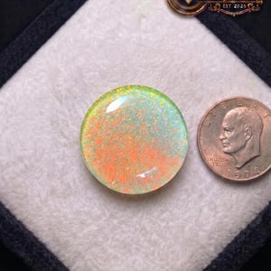 May include: A round, iridescent opal gemstone with orange and blue hues, next to a US dime coin. The opal is set against a white background within a black square. The image includes the text "SHIFA GEMS & JEWELS EST 2026".