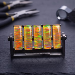 May include: Five rectangular, iridescent orange and green opal gemstones are mounted vertically in a dark metal frame. The base of the frame features a small metal plate with the text SHIFAGEMSANDJEWELS engraved in a clear, sans-serif font.