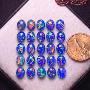 May include: A collection of twenty-five oval-shaped blue opals arranged in a five-by-five grid, each displaying vibrant flashes of red, orange, and green color. A United States one-dollar coin is positioned to the right for scale.