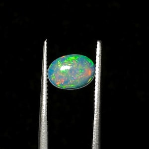 May include: An oval-shaped opal gemstone, held by tweezers against a black background. The opal displays a vibrant play-of-color, with flashes of green, orange, and blue. The gemstone is polished and smooth, reflecting light.