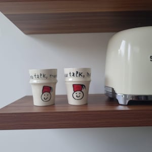 Peut inclure: Deux petites tasses en céramique blanc cassé de forme cylindrique. Chaque tasse présente un visage souriant avec un fez rouge et l'inscription "less talk, more" en lettres noires. Un grille-pain crème est en arrière-plan.