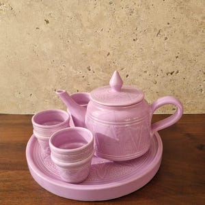 Puede incluir: Un juego de té de cerámica de color lila, que incluye una tetera con tapa, dos tazas pequeñas y una bandeja redonda. La tetera y las tazas tienen un diseño texturizado. El juego se exhibe sobre una superficie de madera.