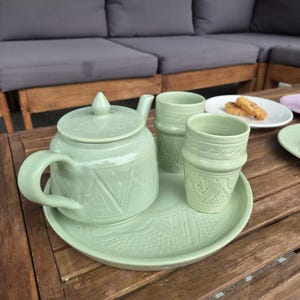 Op de afbeelding: Een licht saliegroene theeset bestaande uit een theepot met deksel, twee bijpassende kopjes en een rond dienblad. Elk stuk is voorzien van een gestructureerd geometrisch patroon met driehoekige en gestippelde motieven in het keramiek.