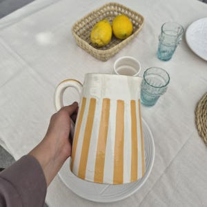 Puede incluir: Una jarra de cerámica con acabado blanco brillante, rayas verticales amarillas y un asa curva. La jarra se sostiene con la mano, mostrando su forma cilíndrica y su textura de superficie lisa, diseñada para servir líquidos en la cocina.