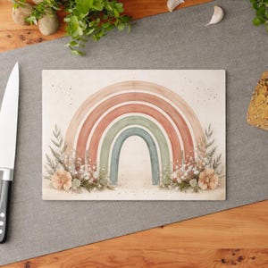 Op de afbeelding: Rechthoekige snijplank met een aquarel regenboogontwerp in perzik-, groen- en blauwtinten. De regenboog is versierd met bloemen en botanische accenten. De plank ligt op een grijze ondergrond.