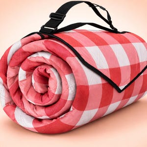Puede incluir: Manta de picnic a cuadros rojos y blancos enrollada, con una correa de transporte negra. La manta está acolchada y tiene un borde negro. Lista para usar al aire libre.