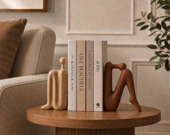 Może przedstawiać: Zestaw czterech książek zatytułowanych The Kinfolk Home, Live Beautiful, A Democracy of Imagery oraz at 100, otoczonych dwiema abstrakcyjnymi podpórkami do książek w kształcie postaci ludzkich, jedna w kolorze beżowym, druga w brązie.