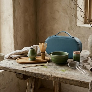 May include: A matcha tea set including a green ceramic bowl, a bamboo whisk, a green whisk holder, a bamboo scoop, a metal strainer, and a small green canister with a copper lid, all arranged with a blue oval carrying case on a wooden surface.