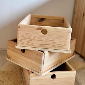 May include: A stack of three rectangular wooden crates made of light-colored pine with visible wood grain. Each crate features a circular cutout handle on the front panel and is constructed with small metal nails along the edges of the side panels.