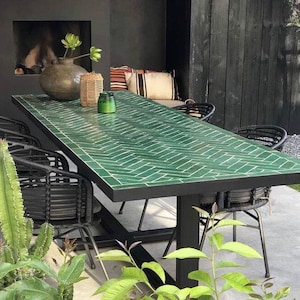 Op de afbeelding: Een rechthoekige eettafel met een groen betegeld oppervlak in een visgraatpatroon met een zwart metalen frame en poten, vergezeld door verschillende zwarte gevlochten stoelen met dunne metalen frames rondom de tafel.