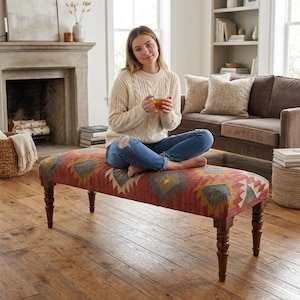 May include: A rectangular upholstered bench featuring a woven fabric top with a red, grey, and cream geometric pattern. The bench is supported by four dark brown turned wooden legs with a classic spindle design, providing a sturdy base.