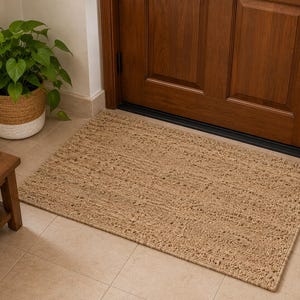 May include: A rectangular doormat with a textured, woven tan surface. The mat features a dense, looped construction made from natural-colored fibers, providing a consistent, neutral appearance that lies flat against the floor in front of a door.