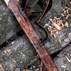 May include: A detailed close-up of a sword in a wooden scabbard with intricate carvings. The sword's hilt is gold-colored with a green gem. A brown leather strap is attached to the scabbard. The scabbard is resting on a bed of wood.