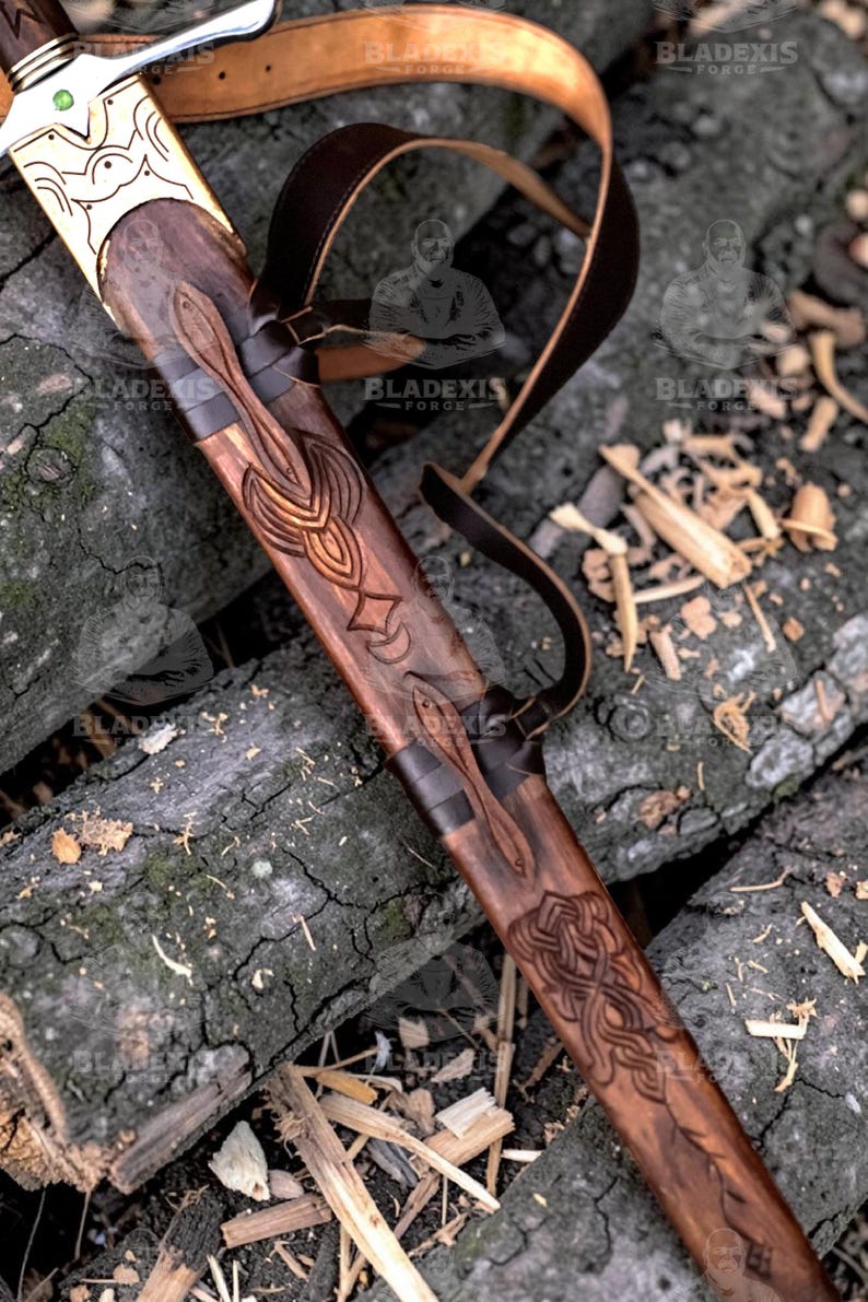 May include: A detailed close-up of a sword in a wooden scabbard with intricate carvings. The sword's hilt is gold-colored with a green gem. A brown leather strap is attached to the scabbard. The scabbard is resting on a bed of wood.