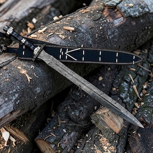 May include: A detailed image of a sword with a patterned blade and a black handle, resting on a pile of wood logs. The sword is in a black sheath with white stitching and the letter "K".
