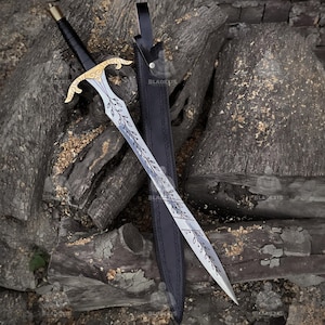 Puede incluir: Una espada plateada con una empuñadura dorada y un mango envuelto en cuero negro descansa junto a su vaina de cuero negro. La hoja presenta un diseño de hojas. La espada se exhibe sobre madera envejecida.