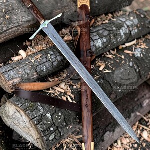 May include: A detailed image of a sword with a patterned blade and a wooden handle. The sword is resting on a pile of logs, with a matching wooden sheath. The handle has intricate carvings and a metal crossguard. The sheath has a leather belt.