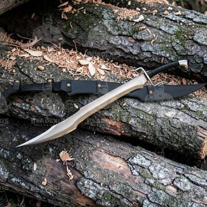 May include: A large, curved sword with a silver blade and black handle rests on a bed of wooden logs. A black leather sheath is behind the sword. The sword has a distinctive, sharp point and a crossguard.