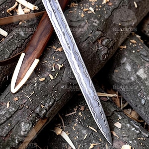 May include: A detailed image of a sword with a patterned blade and a wooden handle. The sword is partially sheathed in a wooden scabbard with a gold-colored tip. The items rest on a bed of wood logs and wood shavings.