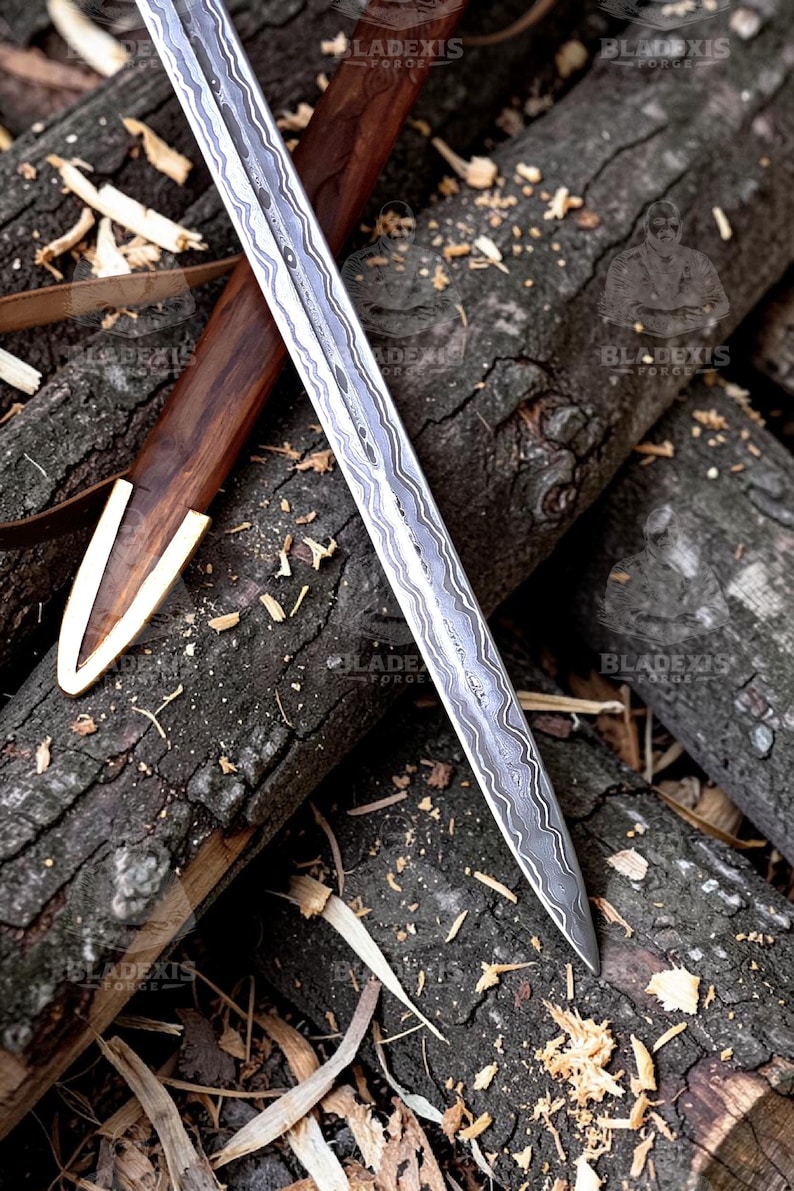 May include: A detailed image of a sword with a patterned blade and a wooden handle. The sword is partially sheathed in a wooden scabbard with a gold-colored tip. The items rest on a bed of wood logs and wood shavings.