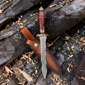 May include: A decorative dagger with a patterned blade and a wooden handle with gold accents. The dagger is in a brown leather sheath, resting on a bed of wooden logs. The handle is a dark red colour.
