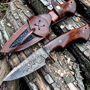 May include: A handcrafted hunting knife with a patterned blade and a wooden handle. The knife is accompanied by a brown leather sheath with decorative stitching and a cross design. The knife and sheath are displayed on a wooden surface.