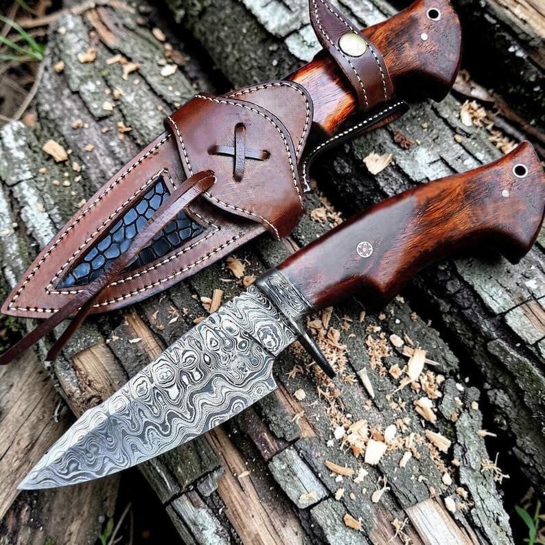 May include: A handcrafted hunting knife with a patterned blade and a wooden handle. The knife is accompanied by a brown leather sheath with decorative stitching and a cross design. The knife and sheath are displayed on a wooden surface.