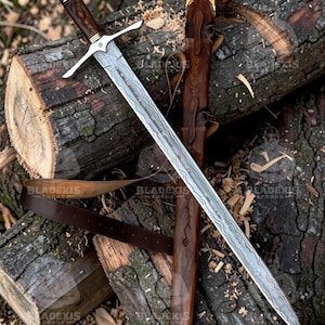 May include: A long, ornate sword with a patterned blade and a wooden handle, resting on a wooden log. The sword has a silver crossguard and a brown leather sheath with gold accents. The blade has a unique pattern.