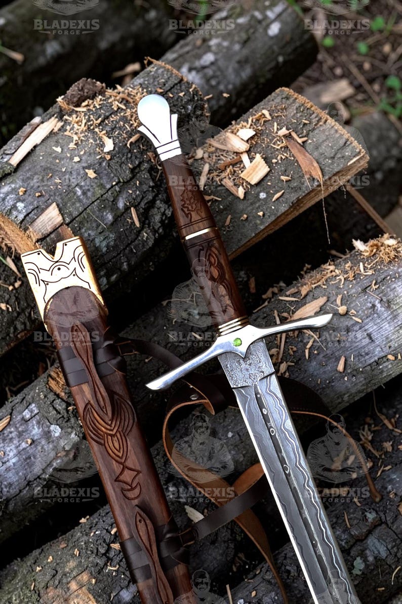 May include: A detailed image of a sword and scabbard. The sword features a silver crossguard and a patterned blade. The handle is dark wood with intricate carvings. The scabbard is also wood, with leather straps and decorative carvings. The sword is resting on a pile of logs.